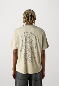 Beige katoenen t-shirt met een lijntekening van een bloemenontwerp en tekst op de achterkant. Korte mouwen, ronde hals en een relaxte pasvorm.
