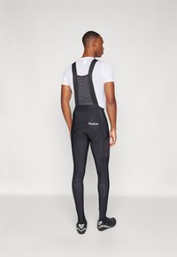 Pantaloni da ciclismo neri con bretelle e schiena in rete, vestibilità aderente e dettagli riflettenti. Indossati con una maglietta aderente bianca e scarpe da ciclismo.
