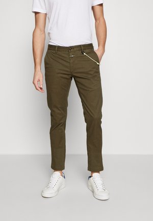 Mann trägt olivgrüne Chinos, ein weißes T-Shirt und weiße Sneaker, steht mit einer Hand in der Tasche vor einem einfarbigen Hintergrund.