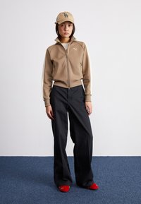 Tan zip-up jacket with white side stripes, black wide-leg pants, and red slippers. Beige cap with LA design and logo. Plain background.