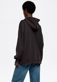 Sudadera grande de color marrón oscuro con capucha con cordón, hombros caídos y puños acanalados, combinada con unos vaqueros anchos de color azul claro.