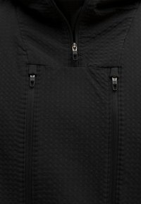 Sort zip-up jakke med tekstureret stof og en prikket mønster. Inkluderer to lynlåslommer på brystet og et højt kravedesign.