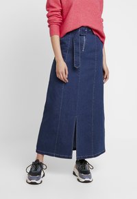 Denim maxikjol i mörkblått med en framknäppning, bälte i midjan och kontrasterande sömmar. Kombinerad med grå och marinblå sneakers.