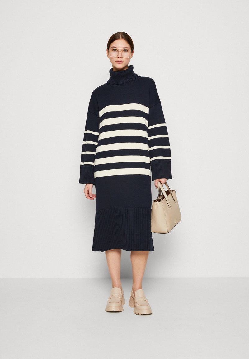 Kobieta ubrana w granatowo-białą w paski sukienkę midi z golfem, beżowe masywne loafersy, przezroczyste okulary i trzymająca beżową torebkę.