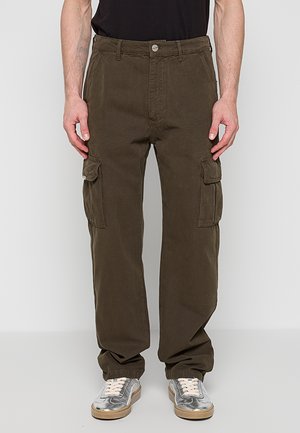 Mężczyzna ubrany w ciemnooliwkowe spodnie typu cargo z bocznymi kieszeniami, czarną koszulę oraz srebrne trampki z białymi sznurowadłami, stojący na białym tle.