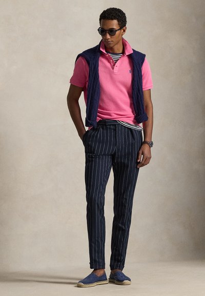 Άνδρας που φοράει σκουρόχρωμα γυαλιά ηλίου, ροζ πόλο πουκάμισο, μπλε πουλόβερ περασμένο στους ώμους, ριγέ μπλε παντελόνια και μπλε παπούτσια χωρίς κορδόνια.