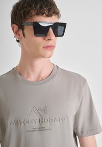 Camiseta de jersey gris con branding en relieve, cuello redondo y mangas cortas. Gafas de sol geométricas negras con lentes oscuros y acentos mínimos.