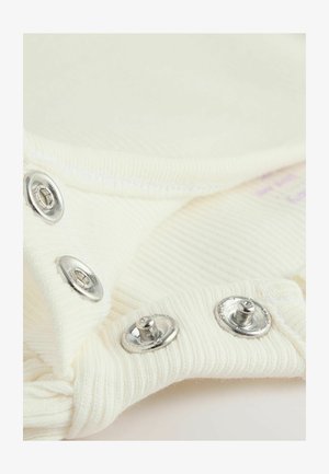 Krém színű, bordázott textúrájú anyag ezüst sznapszerű gombokkal a szélén. Az anyag puhanak és enyhén nyújthatónak tűnik.