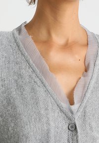 Szary kardigan o miękkiej, dzianinowej fakturze, z dekoltem w kształcie litery V i wykończeniem z surowym brzegiem wzdłuż linii szyi. Posiada zapięcie na guziki z przodu.