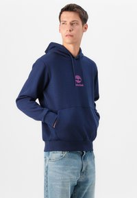 Navy blue hoodie with a pink Timberland logo on the chest, featuring a kangaroo pocket and ribbed cuffs and hem. Textured fabric finish.