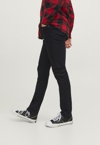 Pantalons noirs à jambe droite avec une texture lisse, portés avec des baskets noir et blanc. La tenue comprend une chemise en flanelle à carreaux rouges et noirs.