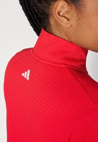 Top desportivo vermelho com colarinho alto, tecido texturizado e detalhe em branco no logo nas costas, apresentando um design ajustado para vestuário ativo.