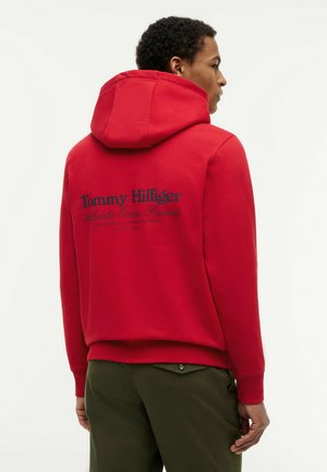 Mężczyzna w czerwonej bluzie z kapturem Tommy Hilfiger z granatowym napisem na plecach i oliwkowych spodniach, stojący tyłem na jednolitym białym tle.