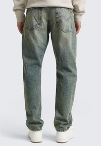 Jeans de mezclilla azul claro con un corte relajado, que incluyen bolsillos traseros, detalles de costura y una textura ligeramente desgastada. Zapatillas blancas visibles.