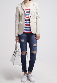 Personne portant un blazer beige par-dessus une chemise rayée rouge, blanche et bleue, un jean foncé déchiré, des baskets blanches, tenant un sac à main blanc sur un fond blanc.
