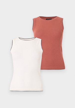 Žena so slnečnými okuliarmi, bezrukávovým hrdzavohnedým topom, širokými modrými nohavicami a béžovými otvorenými lodičkami stojí na bielom pozadí.