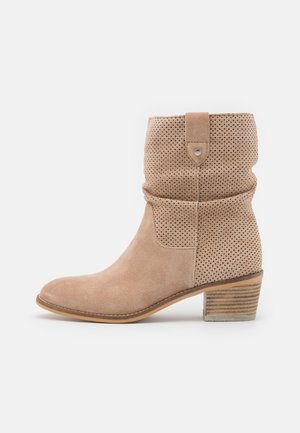 Beige Wildleder-Stiefelette mit perforiertem Obermaterial, Lasche mit Niete und gestapeltem Holzblockabsatz.