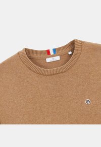 Pull léger couleur marron clair avec col côtelé, petite broche logo ronde sur la poitrine, et rayures rouge, blanc et bleu à l'intérieur du col.