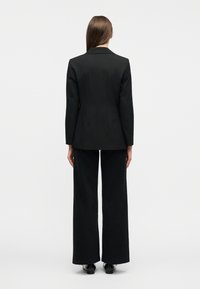 Γυναίκα που στέκεται γυρισμένη με την πλάτη, φοράει μαύρο σακάκι και μαύρο παντελόνι με φαρδύ πόδι πάνω σε απλό ανοιχτό φόντο.