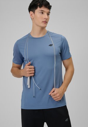 Joven con camiseta deportiva azul y pantalones cortos negros sosteniendo una cuerda para saltar, mirando hacia un lado contra un fondo gris liso.