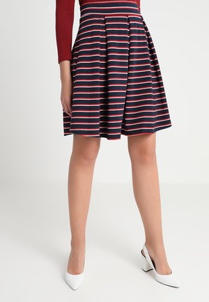 Femme portant une jupe bleu marine à rayures horizontales rouges et blanches ainsi que des escarpins blancs à bride arrière, debout devant un fond uni.