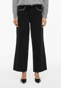 Crne traperice širokih nogavica s zakovicanim detaljima na prednjim džepovima, završene kontrastnim šavovima. Nosi se s ljubičastim pletenim puloverom i crnim cipelama.