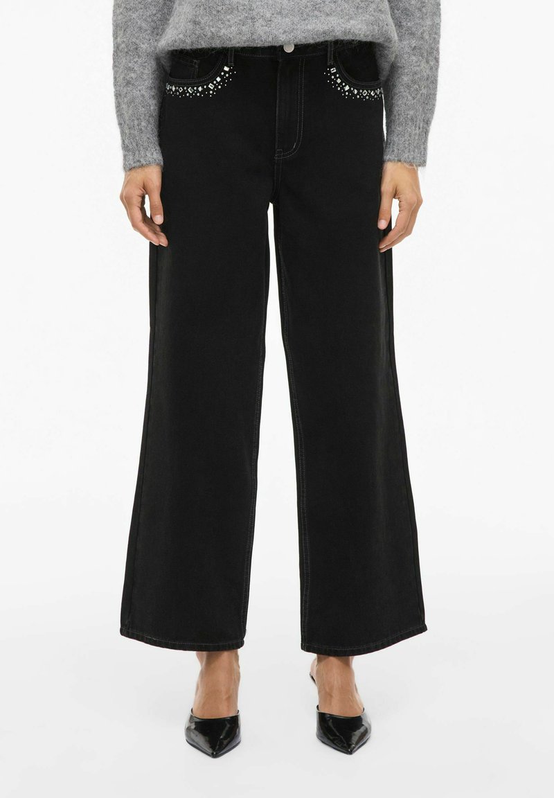 Crne traperice širokih nogavica s zakovicanim detaljima na prednjim džepovima, završene kontrastnim šavovima. Nosi se s ljubičastim pletenim puloverom i crnim cipelama.