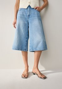 Culottes en denim bleu clair à jambes larges, avec une taille haute et un ourlet brut. Portées avec des tongs noires à imprimé serpent.