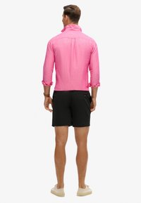 Maglietta lunga rosa, con texture e maniche arrotolate, indossata con pantaloni corti neri e scarpe di colore chiaro, vista di spalle.