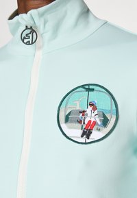 Mentazöld cipzáros dzseki, amelyen egy síelő gőzösben látható, hímzett kör alakú grafika található, fehér cipzárral és lekerekített gallérral.