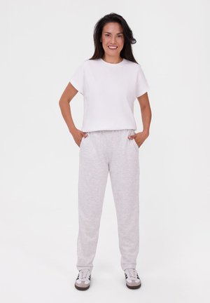 Frau steht mit Händen in den Taschen, trägt ein weißes T-Shirt, hellgraue Jogginghose und weiße Sneakers mit schwarzen Streifen.