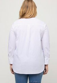 Camicia bianca con bottoni, maniche lunghe, leggera brillantezza, dettaglio della piega posteriore e fessure ai polsini, abbinata a jeans blu. Tessuto dalla trama liscia.