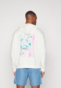 Hvit hettegenser med et blått og rosa abstrakt grafisk design på baksiden, med en teksturert utforming. Brukt sammen med lyseblå shorts.