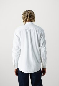 Camisa blanca de manga larga con cuello puntiagudo, puños abotonados y una tela texturizada, combinada con jeans azul oscuro.