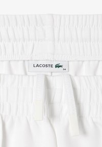 Pantaloni scurți albi cu talie elastică, având etichetă Lacoste cu logo-ul unui crocodil verde și mărimea "34". Material moale, neted, cu o textură verticală fină.
