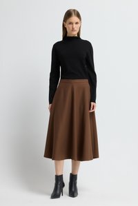 Frau, die ein schwarzes Langarmoberteil, einen braunen Midirock und schwarze Stiefeletten trägt, steht vor einem schlichten hellen Hintergrund.