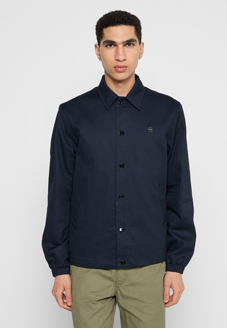 Marineblauwe jas met een kraag, knoopsluiting en elastische manchetten. Soepele stof met twee zijzakken en een klein logo detail.