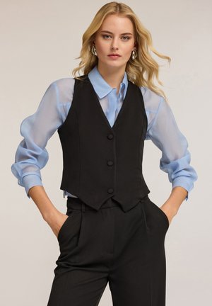 Zwarte op maat gemaakte vest met drie knopen, gedragen over een lichtblauwe doorzichtige blouse met knoopmanchetten. De outfit heeft een getailleerd ontwerp en een gladde textuur.