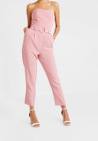 Combinaison rose sans bretelles avec un corsage ajusté, une large ceinture agrémentée d'une boucle circulaire, et un pantalon fuselé longueur cheville. Texture de tissu lisse.