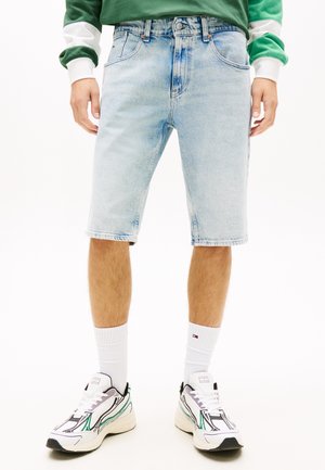 Homme portant un short en jean bleu clair, des chaussettes hautes blanches, des baskets blanches et un sweat-shirt vert avec des rayures blanches sur les manches.