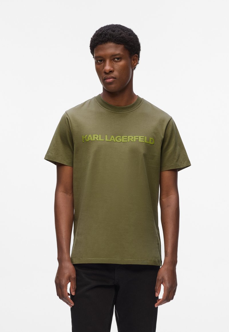 Alyvuogių žalios spalvos medvilnės marškinėliai su apvaliu kaklu, trumpomis rankovėmis ir šviesiai žaliu "KARL LAGERFELD" logotipu atspausdintu per krūtinę.