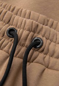 Ceinture en tissu beige avec élastique froncé et deux cordons noirs passés dans des œillets métalliques noirs.