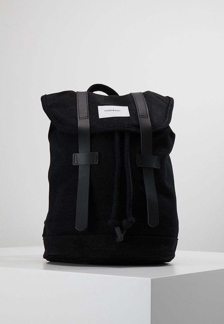 Sac à dos en toile noire avec fermeture par cordon, deux sangles en cuir et une étiquette blanche Sandqvist sur le rabat supérieur, posé sur une surface blanche.