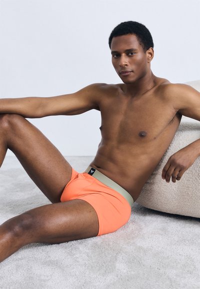 Cuecas boxer laranja com uma cintura cinza claro contrastante. Tecido suave com um ajuste justo, exibido em um modelo numa pose descontraída.