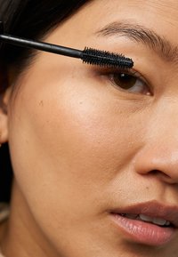 Brosse de mascara noir avec des poils appliqués sur des cils, devant un gros plan d'un teint lisse et clair avec un maquillage subtil et des traits naturels.