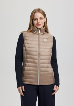 Frau trägt eine beige gesteppte, ärmellose Weste mit Reißverschluss über einem schwarzen Langarmshirt, steht vor einem schlichten grauen Hintergrund.