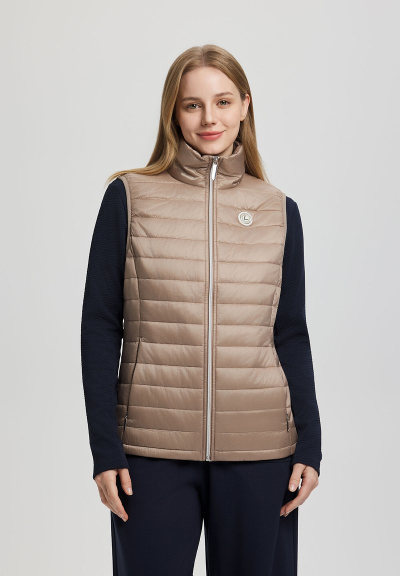 Kvinde iført en beige quiltet, lynlåslukket, ærmeløs vest over en sort, langærmet top, stående mod en ensfarvet grå baggrund.