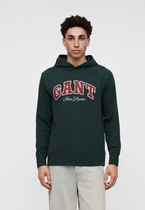 Jeune homme aux cheveux bouclés portant un sweat à capuche GANT New Haven vert foncé et un jean clair, debout devant un fond uni clair.