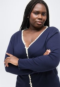 Αυτοπεποίθηνη γυναίκα με μακριά πλεξούδα, ντυμένη με μπλε σκούρο ζακέτα με κρεμ βολάν, με σταυρωμένα τα χέρια μπροστά σε απλό λευκό φόντο.