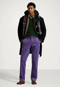 Νεαρός άνδρας με φθινοπωρινό ντύσιμο σε στρώσεις, φορώντας μαύρο παλτό, καρό σκάφος, πράσινο πουλόβερ, ριγέ πουκάμισο, μοβ παντελόνι από κόρντουροι και καφέ παπούτσια.